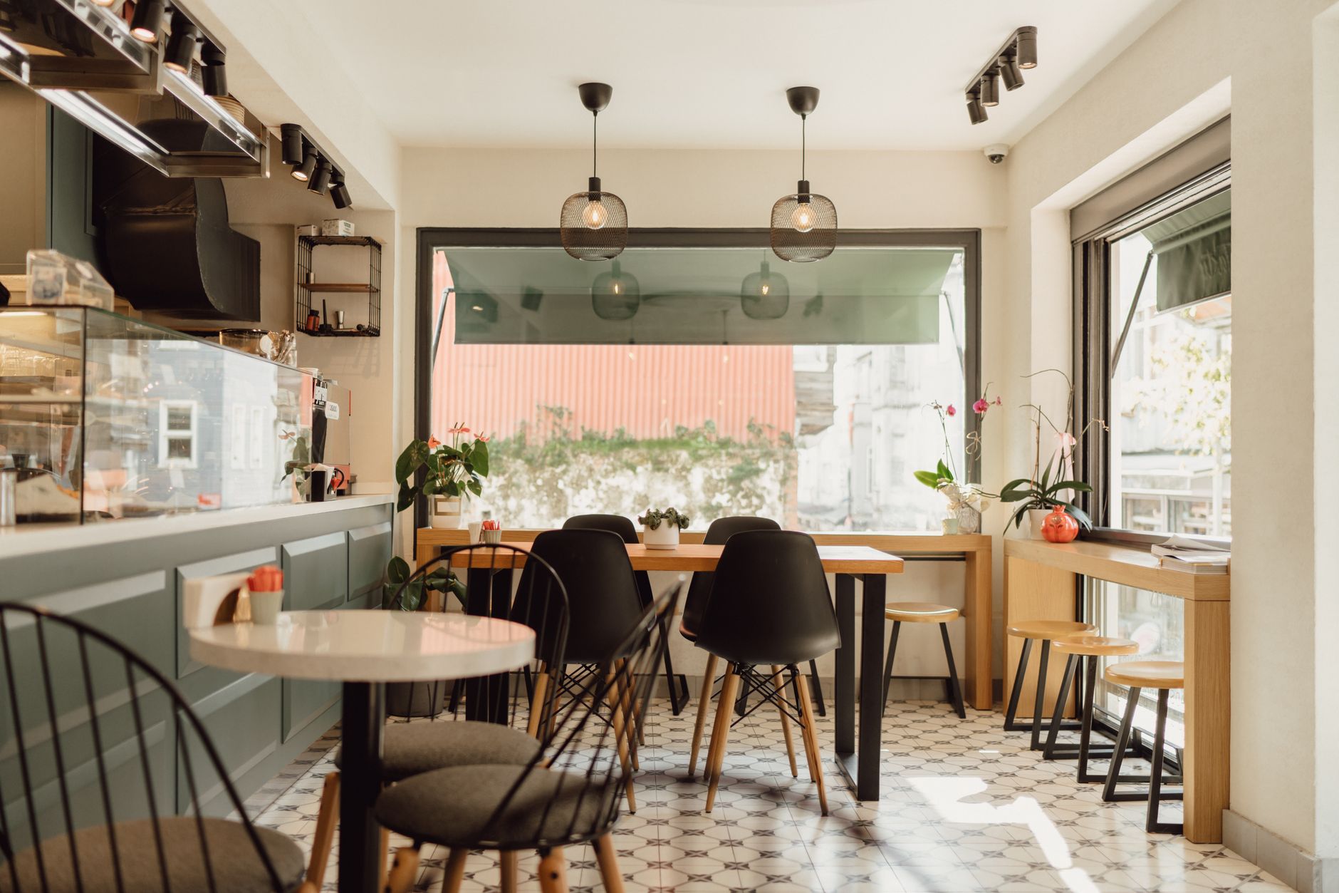 Cafenea goală, cu tejghea, mese și scaune negre lângă o fereastră mare, fără clienți.